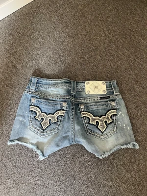 Blå jeansshorts med broderade fickor - Snygga blå jeansshorts från Miss Me med slitna detaljer och råa kanter. Baksidan har broderade fickor med coolt mönster och dekorativa nitar. Perfekta för en avslappnad och trendig look.