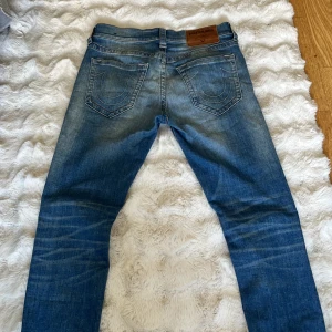 Blå jeans från True Religion - Snygga blå jeans från True Religion med en klassisk femficksdesign och märkets ikoniska logga på bakfickan. Jeansen har en lätt tvättad look och är perfekta för en avslappnad stil. De har en knappgylf och bälteshällor. 