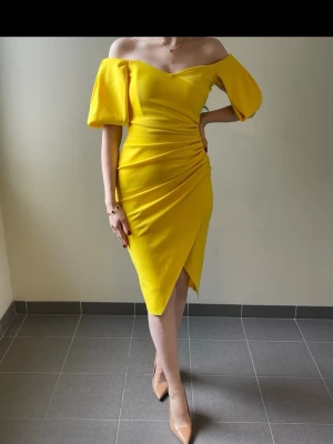Gul offshoulder klänning - Trendig gul klänning med offshoulder och puffiga korta ärmar. Klänningen har omlott-detalj och drapering i midjan som ger en snygg siluett. Perfekt för dig som vill sticka ut med färg och stil.