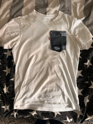 Vit t-shirt med jeansficka - Vit t-shirt med en unik bröstficka i jeans och rutigt tyg. Fickan har råa kanter och en liten röd etikett på sidan. T-shirten har rund hals och korta ärmar, perfekt för en avslappnad stil.