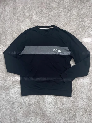 Svart sweatshirt från BOSS - Snygg svart sweatshirt från BOSS med en grå horisontell panel över bröstet och vit BOSS-logga. Tröjan har rund halsringning och långa ärmar. Perfekt för en clean och stilren look.