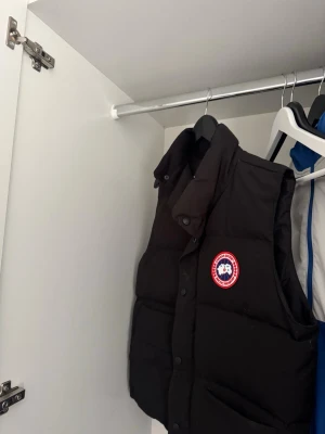 Svart dunväst från Canada Goose - Snygg svart dunväst från Canada Goose med klassisk logga på bröstet. Västen har hög krage, tryckknappar och två stora fickor framtill. Perfekt för lager-på-lager och håller dig varm under kyliga dagar. Äkta väst kom privat 