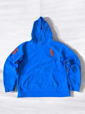 Blå hoodie från Polo Ralph Lauren - Snygg blå hoodie från Polo Ralph Lauren med stor orange broderad logga på bröstet och siffran 3 på ärmen. Klassisk känguruficka framtill och huva. Tillverkad i mjuk bomull som känns skön mot huden. Perfekt för en avslappnad och sportig stil.