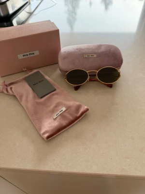 Runda solglasögon från Miu Miu - Snygga runda solglasögon från Miu Miu med guldfärgade metallbågar och mörka linser. Kommer med original rosa sammetsetui, tygpåse och ask. Perfekt accessoar för att lyfta din stil. Går att diskutera priset 