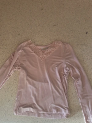Ljusrosa långärmad topp med spets - Söt ljusrosa långärmad topp med v-ringning och dekorativ spetsdetalj runt halsen. Tillverkad i ett mjukt och stretchigt material som känns skönt mot huden. Perfekt för en stilren och feminin look.