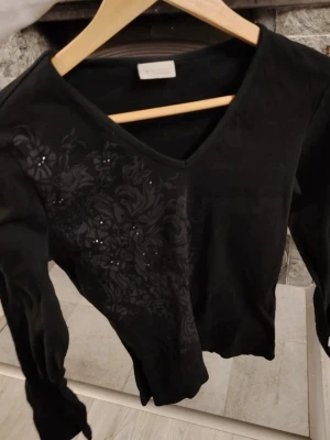 Svart vintage blus med blommönster och V-ringning❤️  - Svart blus från Bon'A Parte med V-ringning och långa ärmar. Ena sidan framtill har ett diskret blommönster och små glittriga detaljer som ger en snygg touch🌸. Perfekt för dig som gillar stilrena plagg med det lilla extra❤️💎 