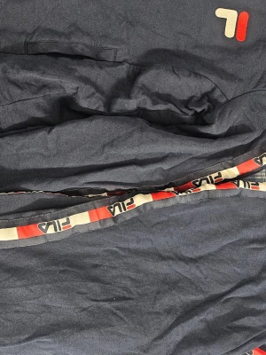 Mörkblå mjukisbyxor från Fila - Mörkblå mjukisbyxor från Fila med vit och röd logga på benet samt Fila-band längs sidorna. Byxorna har en avslappnad passform och är tillverkade i mjukt material, perfekta för chill dagar. Snygg detalj med fickor och elastisk midja.