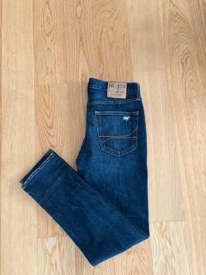 Fadade Straight Fit Hollister Jeans - Snygga mörkblå straight jeans från Hollister med klassisk femficksdesign och schyssta slitningar framtill. Jeansen har knappgylf och en liten logga på bakfickan. Perfekta för en avslappnad och skön look. Storlek W33 L32. Pris kan alltid diskuteras✅