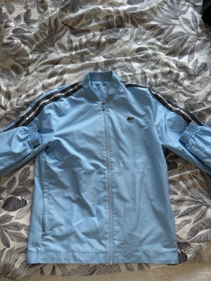 Ljusblått tracksuit-set från Lacoste - Säljer ett ljusblått tracksuit-set från Lacoste med dragkedja på jackan och resår i midja och benslut på byxorna. Setet har svarta och vita detaljer längs ärmarna och byxbenen samt Lacoste-logga på bröstet och byxan. Materialet är troligtvis polyester eller bomullsblandning. Priset kan diskuteras!