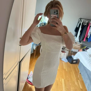 Vit kort offshoulder klänning. Student klänning - Trendig vit kortklänning med puffiga offshoulder-ärmar och figurnära passform. Klänningen har en lätt struktur i tyget och ger en romantisk vibe. Perfekt för sommarens alla tillfällen. Lapp kvar! 