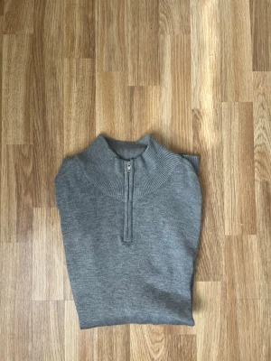 Grå stickad halvzip tröja - Snygg grå stickad tröja med halvzip och hög krage. Tröjan har en stilren design och är tillverkad i mjukt stickat material som passar perfekt till lager på lager. Enkel att matcha med olika outfits.