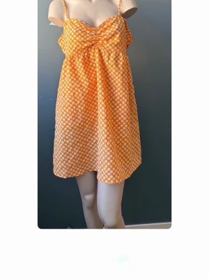 Orange blommig sommarklänning - Söt gul klänning med vita små blommor och tunna axelband. Klänningen har en lätt A-linjeform och är perfekt för varma dagar. Materialet känns lätt och luftigt, vilket gör den skön att bära under sommaren.