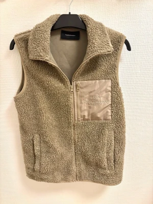 Beige fleeceväst Peak Performance - Mysig beige fleeceväst från Peak Performance med dragkedja framtill och en bröstficka i blankt tyg med broderad logga. Västen har hög krage och två sidofickor, perfekt för lager-på-lager-stil. Materialet är fluffigt teddyfleece.