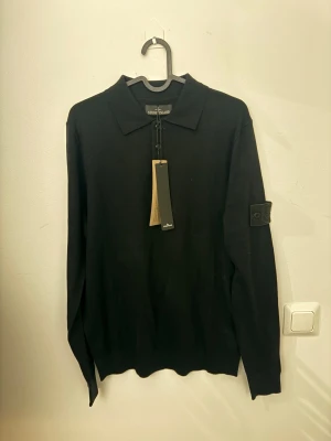 Svart stickad tröja från Stone Island - Svart stickad tröja från Stone Island med klassisk krage och knäppning framtill. Tröjan har lång ärm och ikonisk logotyp på vänster ärm. Perfekt för dig som gillar stilrena och tidlösa plagg.