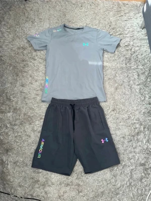 Svarta shorts från Under Armour - Svarta shorts från Under Armour med färgglad logga och texten 'ARMOUR' i regnbågsfärger på vänster ben. Shortsen har elastisk midja med snörning och två sidofickor. Perfekta för träning eller chill.