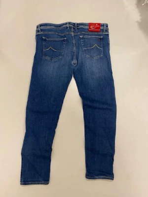 Blå slim fit jeans från Jacob choen  - Snygga blå slim fit jeans från Jacob choen med klassiska fem fickor och diskreta slitningar. Jeansen har en normal midja och raka ben, tillverkade i mjukt denim-material som ger en skön passform. Perfekta för dig som gillar en stilren look.