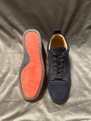 Mörkblå sneakers från Christian Louboutin - Snygga mörkblå sneakers från Christian Louboutin med klassisk röd sula. Skorna har ovandel i mocka, snörning och en stilren design. Perfekta för dig som vill ha något exklusivt och trendigt i garderoben.