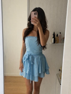 Ljusblå axelbandslös volangklänning - Säljer en ljusblå axelbandslös klänning med smockad överdel och volangkjol i två lager. Klänningen har ett knytband i midjan och är gjord i ett lätt, luftigt tyg som ger en somrig känsla. Perfekt för dig som gillar en söt och enkel stil.