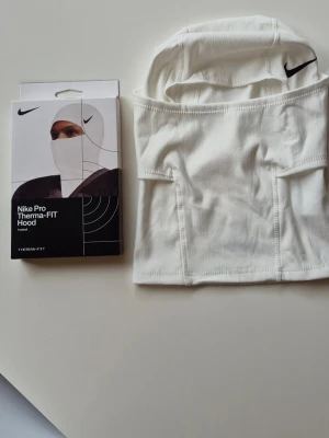 Nike Pro Therma-FIT vit huva - Nike Pro Therma-FIT Hood i vitt, tillverkad i mjukt och stretchigt material. Huvan har en svart Nike-logga framtill och är designad för att ge extra värme och skydd. Perfekt för träning utomhus under kallare dagar.