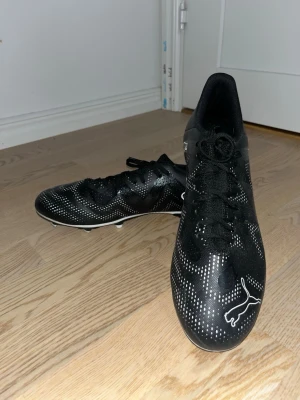 Svarta Puma fotbollsskor med vita detaljer - Säljer ett par svarta fotbollsskor från Puma med vita prickar och logga framtill. Skorna har snörning och låg profil, perfekt för dig som vill ha stilrena och snabba skor på planen. Yttermaterialet är syntet och sulan har dobbar för bra grepp på gräs.