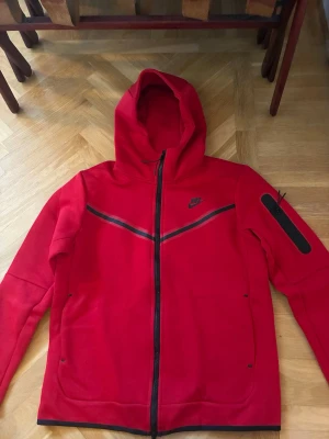Röd Nike hoodie med dragkedja - Cool röd hoodie från Nike med svart dragkedja och detaljer. Har huva och en praktisk ficka med dragkedja på ärmen. Perfekt för dig som gillar sportig stil och vill ha något bekvämt och snyggt. Materialet känns mjukt och skönt mot huden.