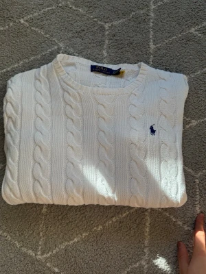 Vit kabelstickad tröja Polo Ralph Lauren - Kabelstickad vit tröja från Polo Ralph Lauren med klassisk blå logga broderad på bröstet. Tröjan har rund halsringning, långa ärmar och ribbade muddar vid ärmslut och nederkant. Perfekt för en clean och stilren look.(Litet hål i mitten som påverkar priset)