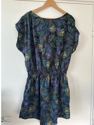 H&M - Kort klänning med påfågelfjädermönster - storlek 42 - Fin glansig mönstrad kort klänning från H&M i storlek 42 (kan även användas som tunika). Svart bas med mönster av påfågel-fjädrar i grönt och lila. Fin klädd knapp vid knäppning bak och bekväma elastiska sömmar i midjan för bästa passform. Mycket gott skick, ny!