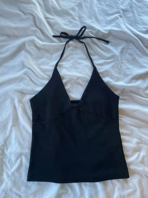 Svart halterneck linnetopp - Snygg svart halterneck linnetopp med djup v-ringning och knyt i nacken. Toppen är ärmlös och har en enkel, stilren design som passar perfekt till sommaren. Tillverkad i mjukt material för en bekväm känsla.