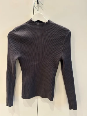 Mörkgrå ribbstickad tröja - Snygg mörkgrå ribbstickad tröja med lång ärm och högre hals. Tröjan har en tight passform och är tillverkad i ett mjukt, stretchigt material som sitter skönt på kroppen. 