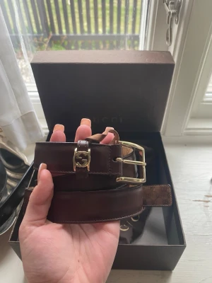 Brunt skärp i skinn från Gucci - Snyggt brunt skärp från Gucci i äkta skinn med guldfärgat GG-spänne och dubbla spännen. Klassisk design med exklusiv känsla, perfekt för att lyfta vilken outfit som helst. Kommer med originalbox och dustbag.