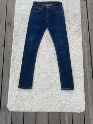 Mörkblå skinny jeans från Nudie - Säljer ett par mörkblå skinny jeans från Nudie med klassiska kontrastsömmar och femficksmodell. Jeansen har en smal passform och är tillverkade i stretchigt denimtyg. Bakfickorna har Nudies ikoniska broderi. Perfekta för en clean och stilren look.