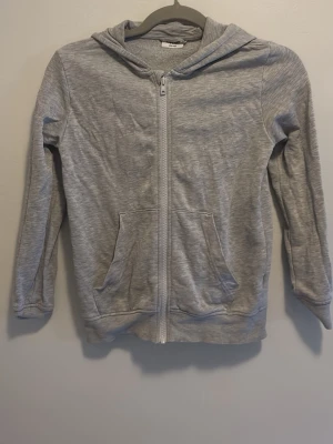 Ljusgrå hoodie med dragkedja - Ljusgrå hoodie med hel dragkedja och två stora fickor framtill. Tröjan har en klassisk huva och är tillverkad i mjukt material som känns bekvämt. Perfekt för en avslappnad stil och passar bra till jeans eller joggers.