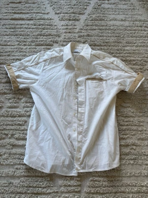 Vit skjorta med beige detaljer - Vit skjorta med korta ärmar och beige detaljer på ärmslut och ficka. Klassisk krage och knäppning framtill. Perfekt för en clean och stilren look.
