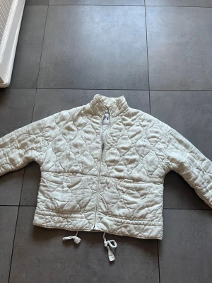 Vit quiltad Zara jacka - Säljer en vit pufferjacka med quiltat mönster och hög krage. Jackan har dragkedja framtill och dragsko i nederkanten för justerbar passform. Perfekt för kalla dagar och riktigt snygg till jeans eller cargo. Materialet känns mjukt och värmande.