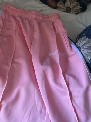 Rosa mjukisshorts från Palm Angels - Säljer ett par snygga rosa mjukisshorts från Palm Angels med vit logga på benet. Byxorna har resår i midjan, sidofickor och vita detaljer längs sidorna. Perfekta för en chill och trendig look.