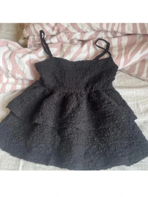 Svart volangtopp med axelband - Supersöt svart topp med tunna axelband och volangdetaljer. Toppen har en strukturerad, lite skrynklig yta som ger en cool look. Perfekt för sommardagar eller när du vill känna dig extra fin.