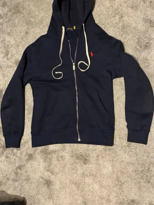Marinblå zip up från Polo Ralph Lauren - Snygg marinblå hoodie med dragkedja från Polo Ralph Lauren. Klassisk design med vit dragsko i huvan och röd broderad logga på bröstet. Mjuk insida och två fickor framtill. Perfekt för en avslappnad och stilren look.