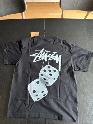 Svart Stüssy t-shirt med tärningsmotiv - Svart t-shirt från Stüssy med stort tryck av två vita tärningar och Stüssy-loggan på ryggen. Framtill finns en mindre logga på bröstet. Klassisk passform och rund halsringning, perfekt för en streetwear-look.