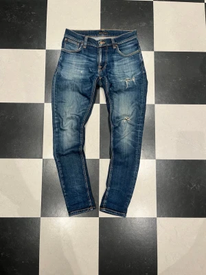 Blå slitna slim jeans från Nudie - Säljer ett par blå skinny jeans från Nudie med slitningar och distressed detaljer på låret och knät. Jeansen har klassisk femficksmodell, orangea sömmar och är tillverkade i stretchig denim för skön passform.