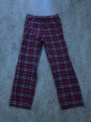 Rödrutiga pyjamasbyxor - Klassiska pyjamasbyxor i rött med vitt och svart rutmönster. Byxorna har rak passform och resår i midjan för extra komfort. Perfekta för mysiga hemmakvällar och gjorda i mjukt tyg som känns skönt mot huden.