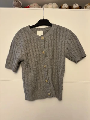 Grå kabelstickad kofta från H&M - Kortärmad grå kabelstickad kofta från H&M med snygga guldfärgade knappar framtill💕