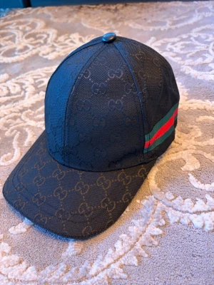 Svart GUCCI keps med gg canvas - Snygg svart keps från Gucci med klassiskt GG-monogram över hela kepsen och en ikonisk grön-röd rand på sidan. Tillverkad i bomull och polyester med elastan för extra komfort. Perfekt accessoar för dig som vill ha en exklusiv och trendig look.
