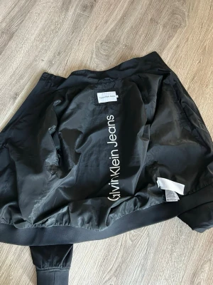 Svart bomberjacka Calvin Klein Jeans - Snygg svart bomberjacka från Calvin Klein Jeans med klassisk passform och ribbade muddar. Jackan har en stilren look med diskret logga på insidan och är tillverkad i ett glansigt syntetmaterial. Perfekt för dig som vill ha en trendig och enkel jacka. Perfekt att ha på våren o sommarn.