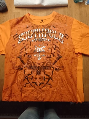Orange Southpole t-shirt med tryck - Snygg orange t-shirt från Southpole med stort grafiskt tryck och texten 'Truth & Liberty' på framsidan. Klassisk rund hals och korta ärmar. Perfekt för dig som vill sticka ut med en färgstark och unik stil.