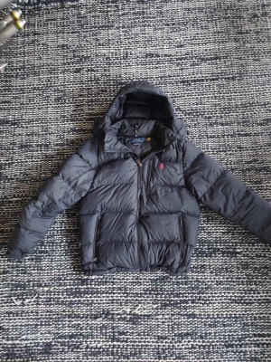 Svart pufferjacka från Polo Ralph Lauren - Snygg svart pufferjacka från Polo Ralph Lauren med huva och dragkedja framtill. Jackan har det klassiska röda Polo-logotypen broderad på bröstet och två sidofickor med dragkedja. Perfekt för kalla vinterdagar och riktigt skön att bära.