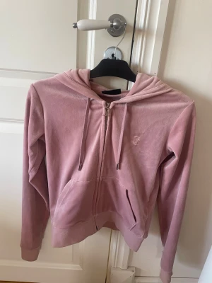 Rosa velourhoodie Juicy Couture - Säljer en ikonisk rosa hoodie från Juicy Couture i mjuk velour. Dragkedja framtill med snygg J-detalj, fickor och huva med dragsko. Perfekt för en chill och trendig look. Broderad logga på bröstet och klassisk passform.