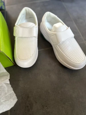 Vita loafers med kardborreband - Snygga vita loafers med perforerat mönster på ovandelen och praktiskt kardborreband. Skorna har en klassisk form och är tillverkade i ett slätt syntetmaterial. Perfekta för dig som gillar stilrena och bekväma skor.