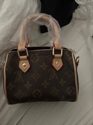  brun monogram handväska - Snygg brun handväska  med klassiskt monogrammönster. Väskan har två korta handtag i beige läder med röda detaljer och guldfärgade metalldelar. Dragkedja upptill och rymligt fack inuti. Perfekt accessoar för dig som gillar lyxig stil.