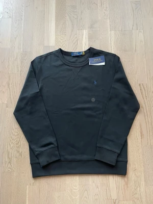 Svart sweatshirt från Polo Ralph Lauren - Stilren crewneck från Polo Ralph Lauren i mörk färg ✨  En tidlös och stilren sweatshirt med klassisk broderad logga på bröstet. Perfekt för både vardag och mer uppklädda outfits. Kommer med original tags.  • Märke: Polo Ralph Lauren   • Typ: Crewneck sweatshirt/Tunn • Färg: Mörkblå / svart   • Passform: Regular fit  M/S • Skick: Ny med tags   • Mjuk och bekväm premiumkvalitet    Lätt att matcha med jeans, chinos eller under en jacka. Ett måste i garderoben.