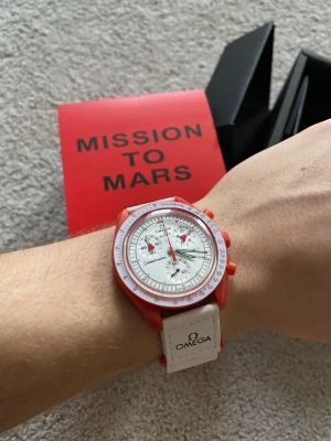 Omega x Swatch Mission to Mars klocka - Säljer en Omega x Swatch Speedmaster Mission to Mars från Bioceramic MoonSwatch-kollektionen. Klockan har röd boett, vit urtavla med röda detaljer och vit rem med Omega-logga. Snygg och unik design som sticker ut på handleden.